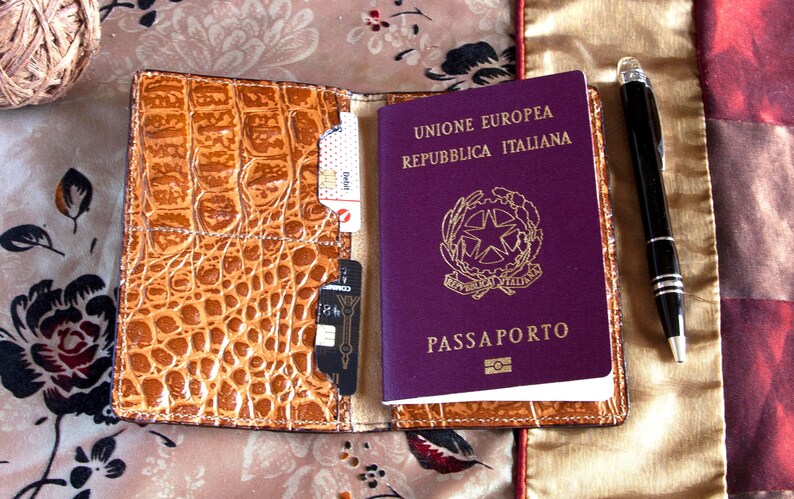 Puede incluir: Un portapasaporte de cuero marr&oacute;n con un pasaporte italiano morado en su interior. El pasaporte tiene el texto "Unione Europea Repubblica Italiana" y "Passaporto" en la portada. Un bol&iacute;grafo plateado est&aacute; sobre una superficie de tela beige junto al portapasaporte.