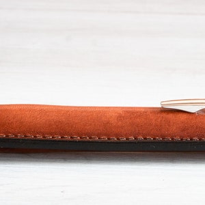 May include: A brown leather pen case with a black trim and a silver pen inside.