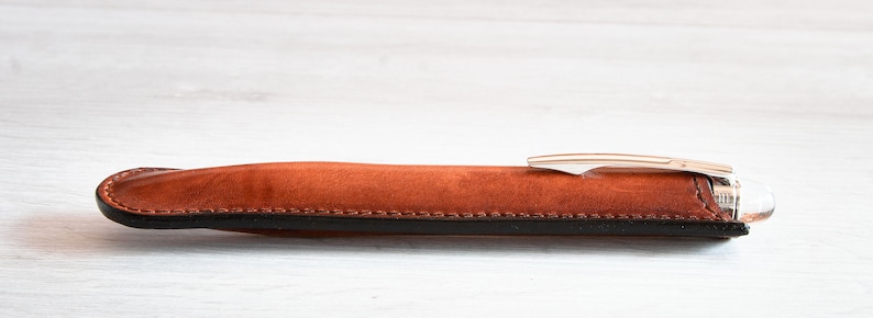 May include: A brown leather pen case with a black trim and a silver pen inside.