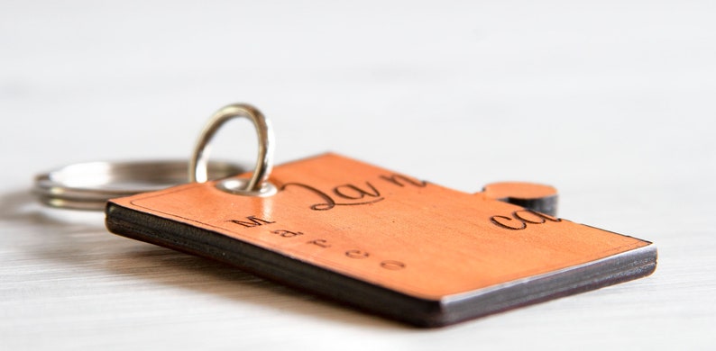 May include: A wooden keyring with a rectangular tag and a small circular accent. The tag is light brown with dark brown edges and features engraved text. A silver metal ring is attached for keys.