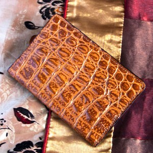 Puede incluir: Una cartera de cuero marr&oacute;n con un patr&oacute;n de cocodrilo. La cartera est&aacute; sobre una tela estampada.