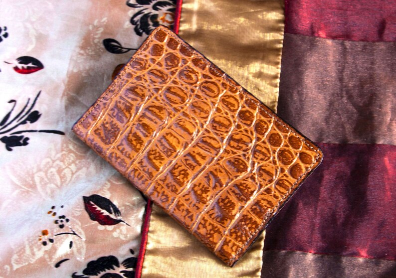 Puede incluir: Una cartera de cuero marr&oacute;n con un patr&oacute;n de cocodrilo. La cartera est&aacute; sobre una tela estampada.