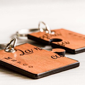 May include: Two wooden puzzle piece keyrings with silver key rings. The keyrings are light brown with black edges and engraved names and text. One keyring says "Marco" and the other says "Laura".