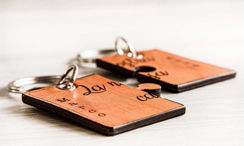 May include: Two wooden puzzle piece keyrings with silver key rings. The keyrings are light brown with black edges and engraved names and text. One keyring says "Marco" and the other says "Laura".