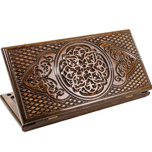 May include: A dark brown, wooden backgammon board with intricate carved floral and geometric patterns. The board is open, revealing the playing surface and is set against a white background. The detailed craftsmanship is evident in the ornate design.
