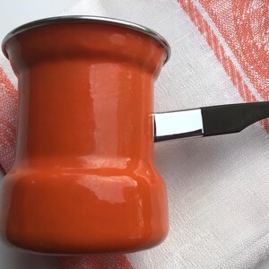 May include: An orange enamel coffee pot with a black handle and a silver rim. The pot is sitting on a white and orange striped kitchen towel. The pot is designed for making coffee.