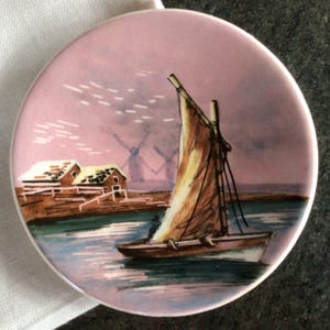 May include: A pink ceramic saucer featuring a painted scene of a sailboat on a body of water with windmills in the background. The scene is painted in shades of brown, yellow, and green.