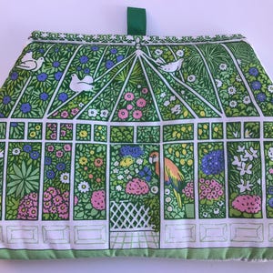 May include: Green and white fabric pot holder with a floral and bird design. The pot holder features a parrot and a greenhouse design.