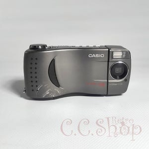 Op de afbeelding: Een grijze Casio digitale camera met een lens, flitser en knoppen. De merknaam "CASIO" staat op de voorkant. De lens is f=3,0 mm 1:2,0. De camera heeft krassen aan de zijkant.