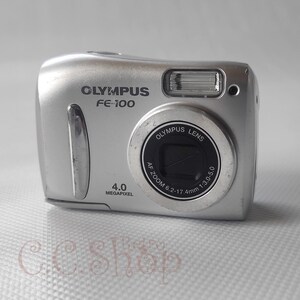 May include: Silver Olympus FE-100 digital camera. The camera has a lens with the text "Olympus Lens" and "AF Zoom 6.2-17.4mm 1:3.0-5.0". It also has "4.0 Megapixel" printed on the front. The camera has a flash and a silver body.