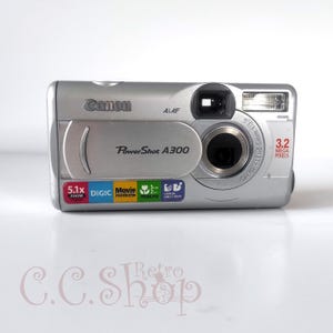 May include: Silver Canon PowerShot A300 digital camera. Features include 5.1x zoom, DIGIC, movie mode, 5cm macro, and 3.2 megapixels. The camera has a built-in flash and a lens with the text "CANON LENS 5.1-15.3mm 1:2.6-4.8".