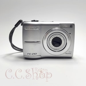 May include: Silver Olympus FE-210 digital camera with a 7.1 megapixel resolution. The camera has a built-in flash and a zoom lens. It includes a grey wrist strap and the Olympus logo is visible on the front.