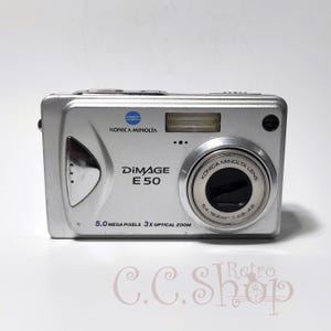 Puede incluir: Cámara digital plateada Konica Minolta DiMAGE E50. Cuenta con una lente con el texto "KONICA MINOLTA LENS" y "5.0 MEGAPIXELS 3X OPTICAL ZOOM". La cámara tiene flash y una pequeña pantalla. Sobre una superficie blanca.