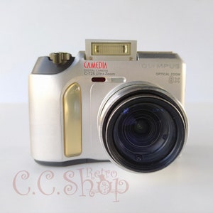 May include: A silver Olympus Camedia C-725 Ultra Zoom digital camera featuring an 8x optical zoom lens, a pop-up flash unit, and a gold-toned vertical accent panel on the front body. The camera body displays the text Camedia, C-725 Ultra Zoom, and 8x.