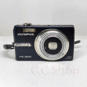 May include: A black Olympus FE-300 digital camera with a silver lens and flash. The camera has the brand name "OLYMPUS" printed on the front. The camera has a 12.0 megapixel resolution.