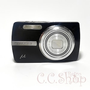 以下が含まれることがあります： 銀色のレンズが付いた黒いオリンパスμデジタルカメラ。レンズには「Olympus Lens AF 5x Optical Zoom 6.4-32mm 1:3.3-5.9」と印刷されています。