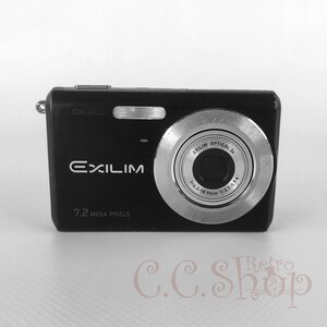 Puede incluir: Cámara digital Casio Exilim negra con lente plateada y el texto "7.2 Mega Pixels". La cámara tiene un pequeño lazo plateado en el lateral. La cámara está sobre una superficie blanca.