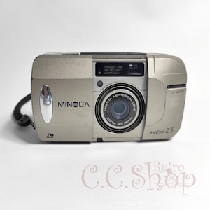 以下が含まれることがあります： ズームレンズ、内蔵フラッシュ、リストストラップを備えたシルバーのミノルタベクティス25カメラ。「MINOLTA」と「VECTIS 25」の文字がプリントされています。レンズには「ZOOM 30-75mm」の文字がプリントされています。