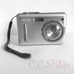 May include: Silver BenQ DC E600 digital camera with a lens and a wrist strap. The camera has a brushed metal finish and the text "BenQ" is visible on the front. The lens has "1X OPTICAL ZOOM 5.8-17.4mm 1:2.8-5.8" printed on it.