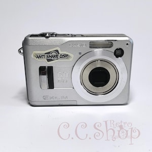 May include: Silver Casio Exilim digital camera with a zoom lens and the text "ANTI SHAKE DSP". The camera has a 6.0-megapixel resolution and a small flash. The camera is a compact size.