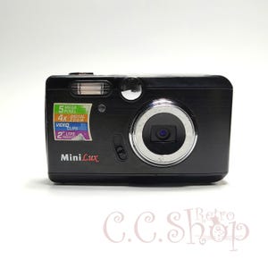 Puede incluir: Cámara digital Mini Lux negra con un anillo de lente y flash plateados. Cuenta con una cámara de 5 megapíxeles, zoom digital 4x, videoclips y una pantalla LTPS de 2 pulgadas. La cámara tiene el texto "Mini Lux".