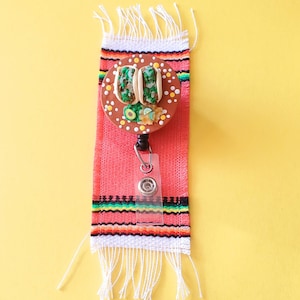 May include: A red and white striped fabric with fringe, featuring a brown and white badge reel with a miniature taco design. The badge reel has a clear plastic badge holder.