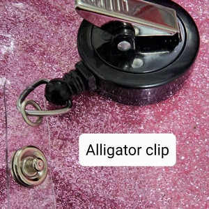 May include: A black retractable badge holder with a silver alligator clip. The holder is on a pink glitter background.