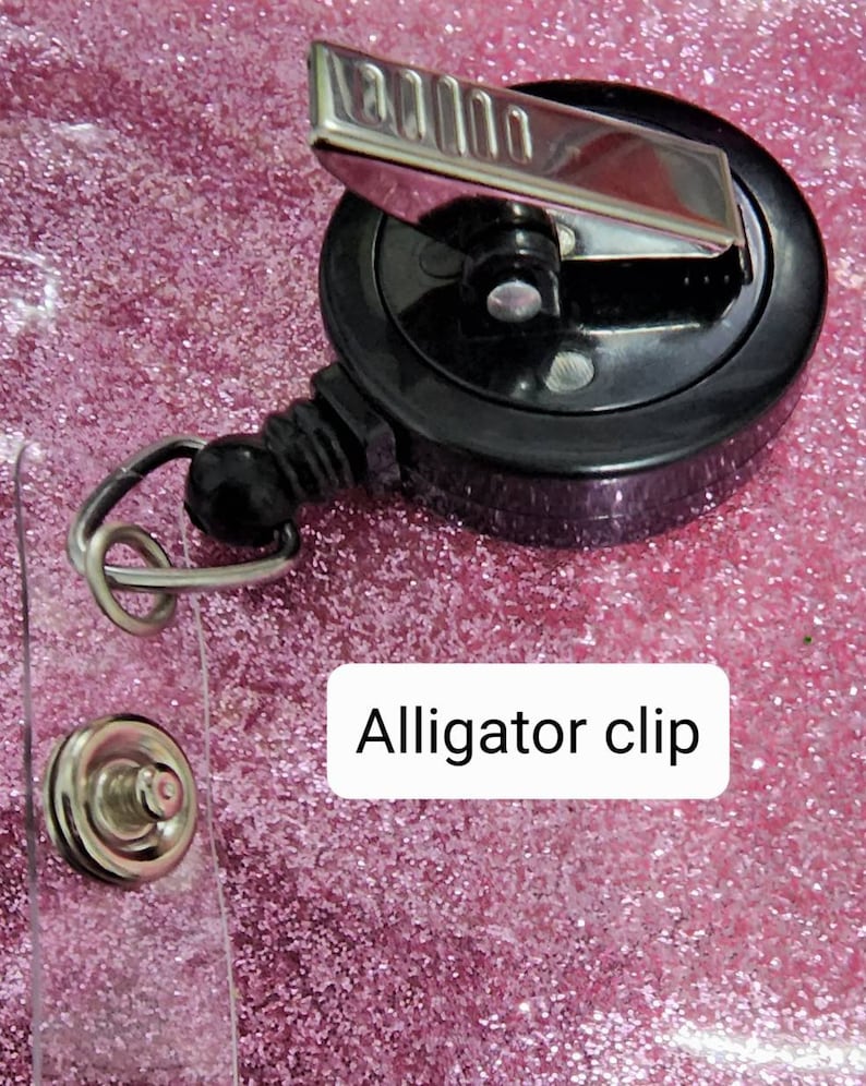 May include: A black retractable badge holder with a silver alligator clip. The holder is on a pink glitter background.