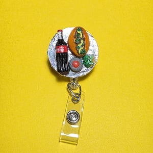 May include: A novelty badge reel featuring miniature food items. The reel includes a small bottle of Coca-Cola, a hot dog, and other food-themed elements. The reel has a clear plastic clip for attaching to clothing or accessories.