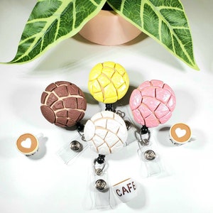 May include: Four colorful, round, bread-shaped charms with a textured surface, attached to retractable badge holders with clear plastic ID card holders. The charms are brown, yellow, pink, and white.  The badge holders also include a small charm of a coffee cup with the word "CAFE" on it.  The text "NEW STYLE" is at the bottom of the image.