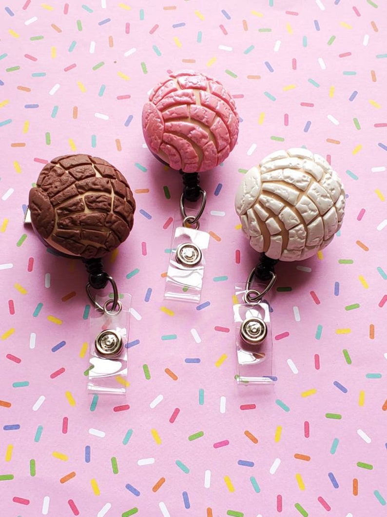 May include: Three colorful, round, pastry-shaped badge holders with a white background. The badge holders are pink and white, brown and white, and white.