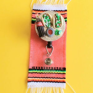 May include: A colorful Mexican-style fabric with a white fringe hangs from a retractable badge holder. The holder features a miniature scene of tacos, a lime wedge, salsa, and a Coca-Cola bottle.