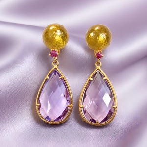 May include: A pair of gold-coloured drop earrings. Each earring features a faceted, teardrop-shaped, light purple gemstone framed in gold. A small, round, red gemstone connects the teardrop to a textured, gold-coloured sphere at the top.