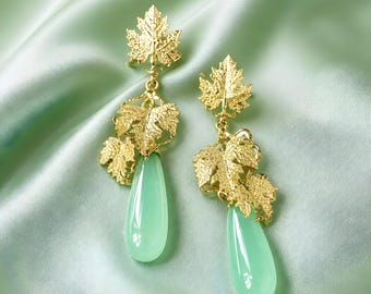 Op de afbeelding: Een paar goudkleurige oorbellen met een bladeren ontwerp. De oorbellen hebben een cluster van gouden bladeren en een druppelvormige, lichtgroene steen. De oorbellen worden gepresenteerd op een lichtgroene satijnen stof.