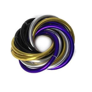 Op de afbeelding: Een verzameling kleurrijke armbanden in goud, zilver, zwart en paars, gerangschikt in een cirkelvormig patroon. De armbanden zijn gemaakt van een glanzend materiaal en zijn op elkaar gestapeld. De achtergrond is effen wit.