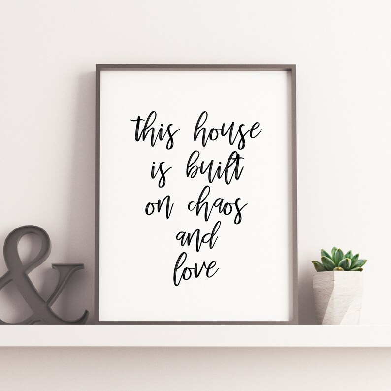 May include: Black and white typography print with the text "This house is built on chaos and love" in a modern calligraphy style. The print is framed and displayed on a white shelf with a small succulent plant in a gray pot.
