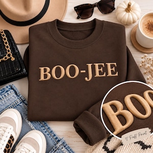 Könnte beinhalten: Ein dunkelbraunes Sweatshirt mit Rundhalsausschnitt und dem Text BOO-JEE in erhabenem, hellbraunem Puff-Druck. Das Sweatshirt hat lange Ärmel und einen gerippten Ausschnitt, mit einem kreisförmigen Einsatz, der eine Nahaufnahme der strukturierten Buchstaben zeigt.