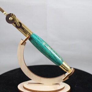 Könnte beinhalten: Ein tealfarbener und goldener Stift mit einem Messingmechanismus und einem Holzständer. Der Stift ist aus Holz mit einer tealfarbenen und goldenen Oberfläche.