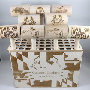 May include: A wooden stand with a Maryland flag design holds a variety of wooden mallets. The mallets are engraved with different designs, including a crab, a state outline, and a sports team logo. The stand has a sign that reads "Custom Designs Available" and "$5 ea 5 for $20".