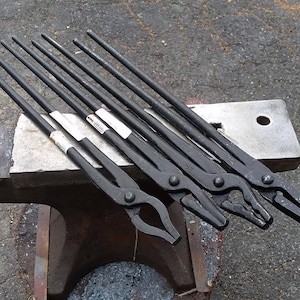 May include: A set of seven black metal blacksmith tongs, resting on a metal anvil. The tongs are of varying lengths and have different jaw shapes.