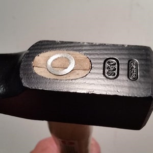 Pode incluir: Close-up de uma cabeça de martelo preta com cabo de madeira. A cabeça do martelo tem uma forma retangular com uma face de impacto plana. A cabeça tem uma inserção de metal circular e marcações estampadas. O cabo é castanho claro.