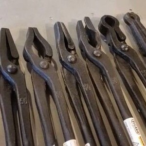 May include: A set of nine black metal blacksmith tongs with various sizes and shapes. The tongs are arranged in a row on a gray surface. The tongs are used for holding and manipulating hot metal.