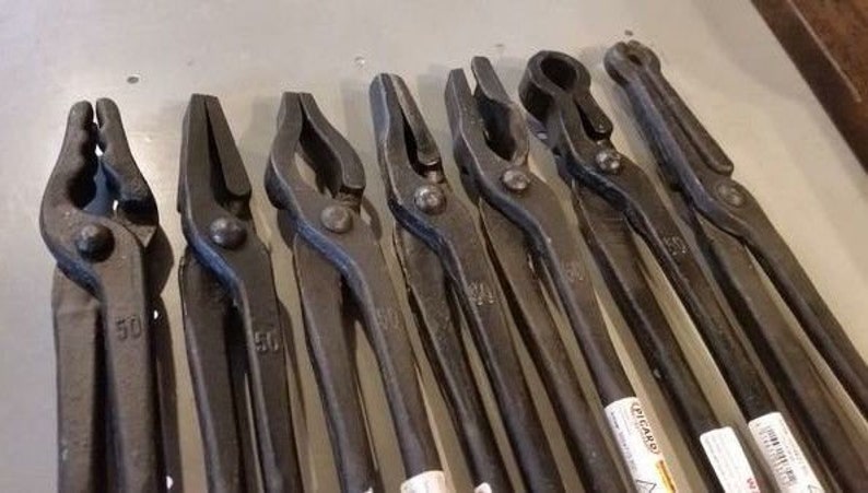 May include: A set of nine black metal blacksmith tongs with various sizes and shapes. The tongs are arranged in a row on a gray surface. The tongs are used for holding and manipulating hot metal.