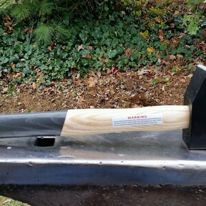 Könnte beinhalten: Ein schwarzer Hammer mit einem Holzstiel und einem schwarzen Gummigriff. Der Hammer liegt auf einem Amboss aus Metall. Der Stiel hat ein Warnschild.