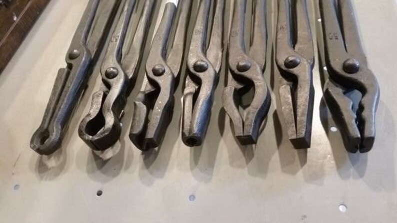 May include: A set of ten blacksmith tongs with various jaw shapes and sizes. The tongs are made of metal and have a dark gray finish.