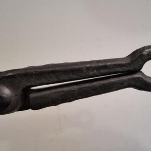 May include: Black metal blacksmith tongs with a rounded handle. The tongs are used to hold hot metal.