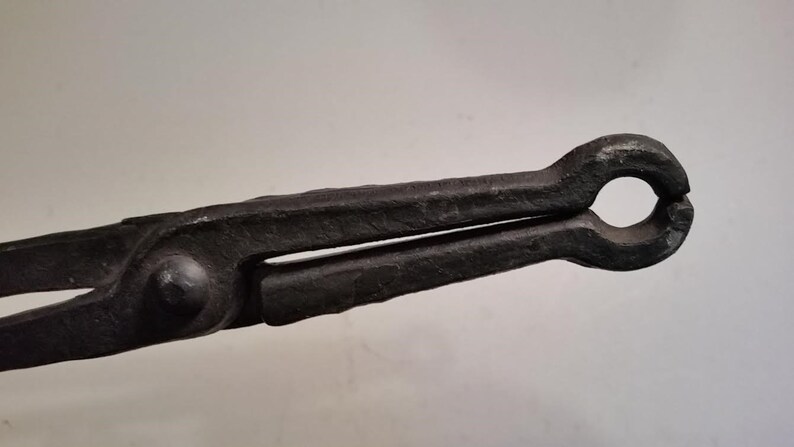 May include: Black metal blacksmith tongs with a rounded handle. The tongs are used to hold hot metal.