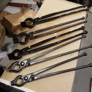 Puede incluir: Cinco tenazas de herrero de metal negro con extremos redondeados. Las tenazas son de diferentes tamaños y están dispuestas en una fila sobre una superficie de madera marrón claro.