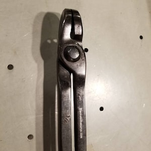 May include: Black blacksmith tongs with a 1/2 inch marking. The tongs are made of metal and have a black finish. The text "Blacksmithsource.com" is engraved on the side of the tongs.