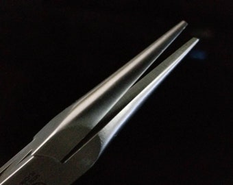 May include: Close-up of needle-nose pliers with a silver finish. The pliers are open, revealing the pointed tips. The tool is set against a dark background, highlighting the metallic sheen and precision of the instrument.